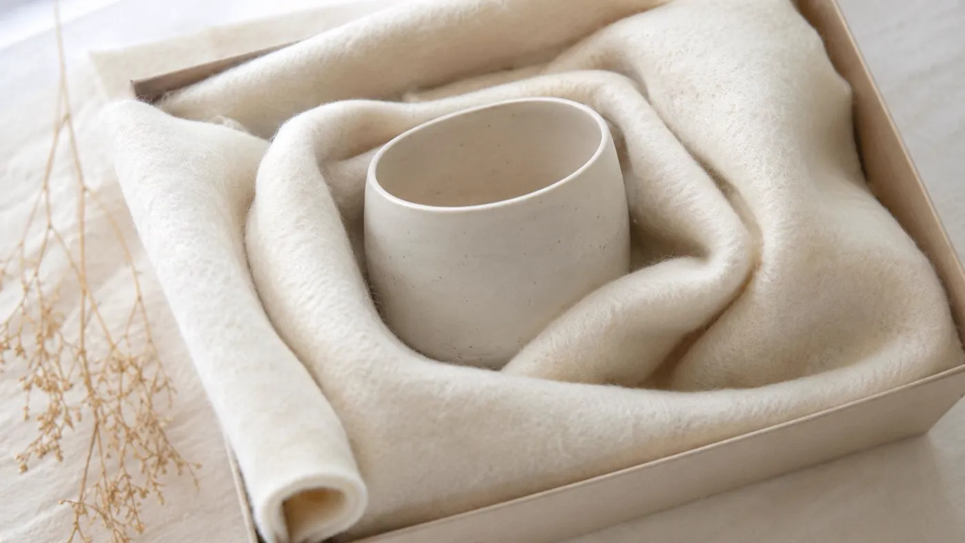 A minimalist white ceramic cup nestled in a soft, fluffy off-white blanket inside a gift box.